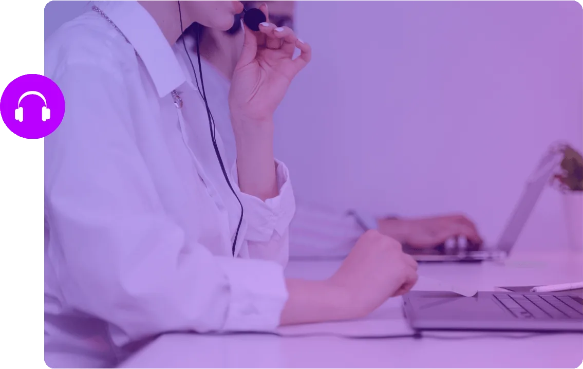Técnica de apoio ao cliente com auscultadores e microfone, ao computador, com um colega ao fundo.