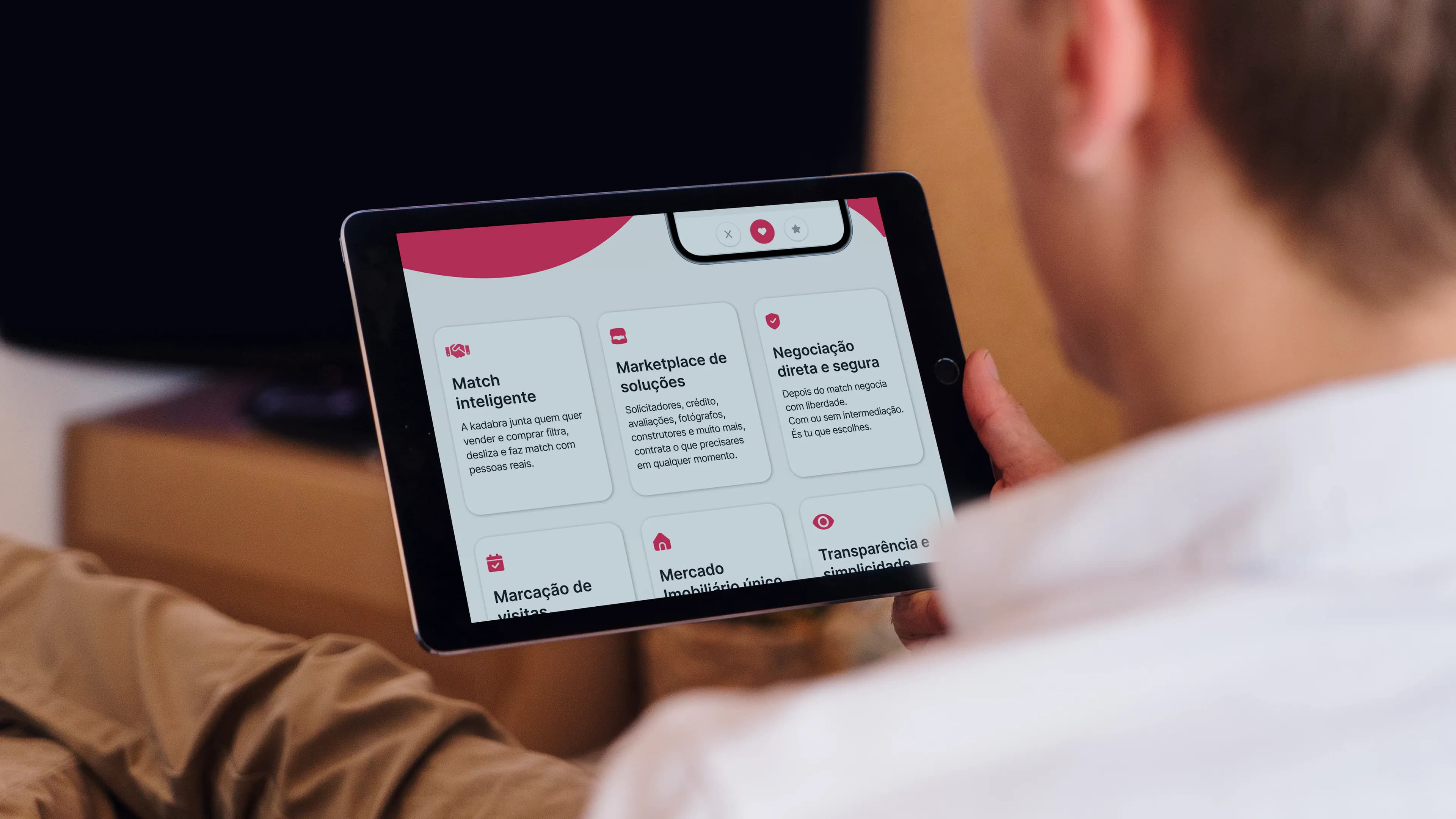 Pessoa a segurar um tablet com a aplicação Kadabra aberta num ecrã de cartões, cada um com ícone e descrição de uma funcionalidade.