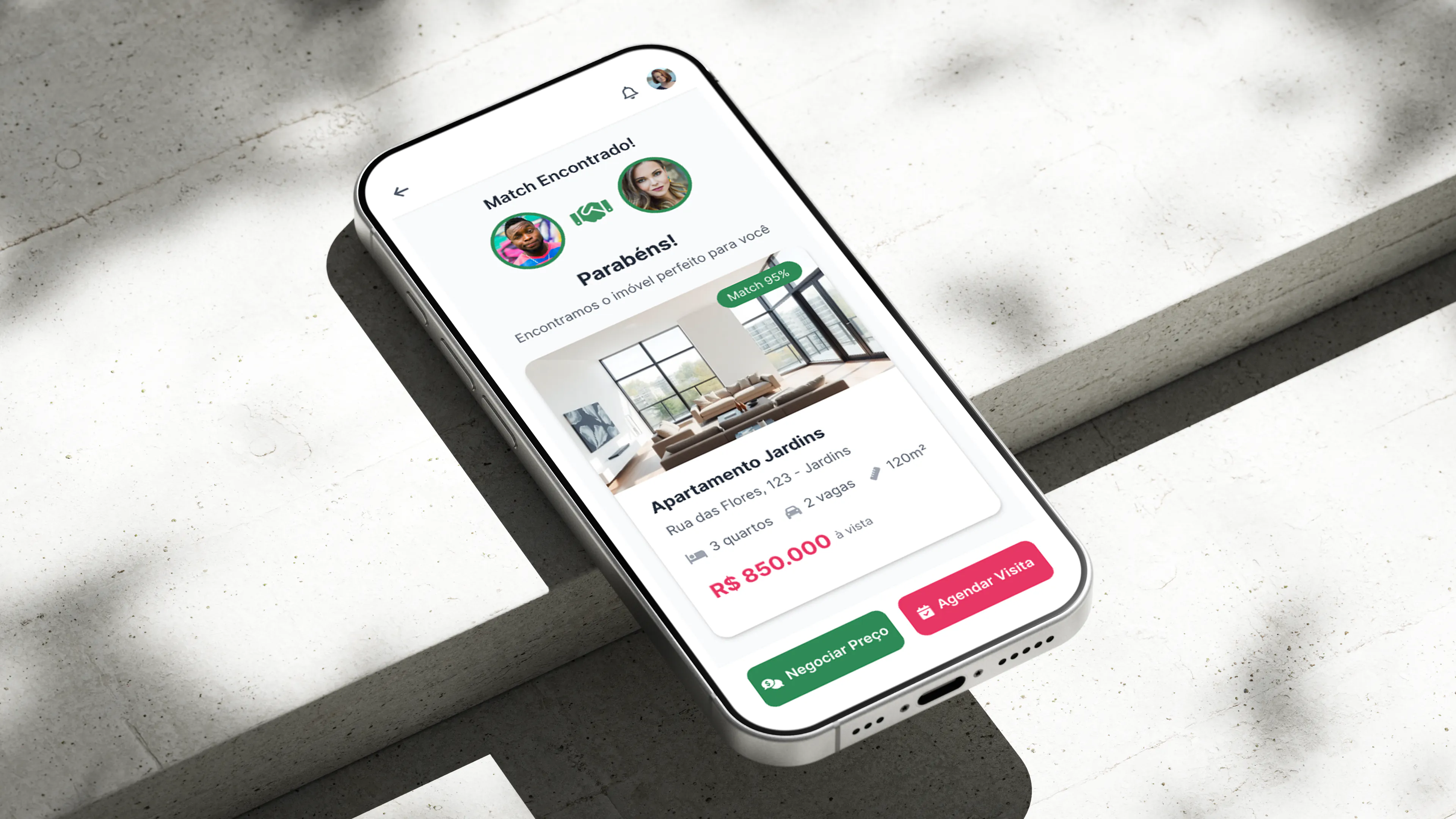 Telefone assente numa superfície de betão, em ângulo, com o ecrã de match da aplicação Kadabra a mostrar a ficha de um apartamento com fotografia, área, número de quartos, preço e botões de negociação e de marcação de visita.